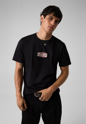 Mand iført en sort Guess Jeans t-shirt, sorte bukser, sølvhalskæde og ringe, stående mod en ensfarvet grå baggrund.