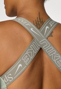 Sportski grudnjak s trakama u obliku slova X, maslinaste boje, s bijelim Nike logom. Mekana tekstura s glatkom finišom i minimalnim hardverom.