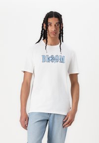 Hvit bomullst-skjorte med en blå "BLOOM" grafikk med blomsterdetaljer. Korte ermer, rund hals, avslappet passform, kombinert med lyseblå jeans.