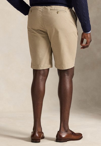 Άνδρας που φοράει μπεζ σορτς, μπλε πουλόβερ και καφέ δερμάτινα μοκασίνια, στέκεται με την πλάτη προς την κάμερα σε ουδέτερο φόντο.