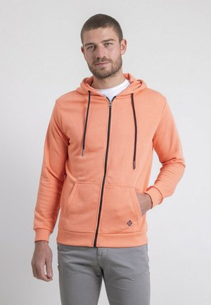 Homme portant un sweat à capuche zippé pêche avec des cordons noirs et un pantalon gris, debout devant un fond blanc uni.