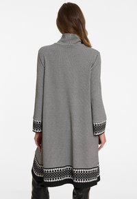 Schwarzer und weißer Hahnentrittmuster langer Mantel mit Kragen, kontrastierenden gemusterten Manschetten und dekorativem Besatz entlang des Saums.