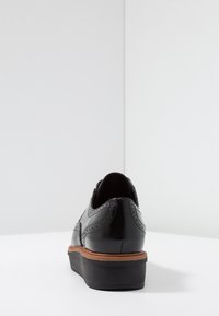 Chaussure en cuir noir avec un design brogue, présentant une tige lisse, des accents texturés, et un talon marron sur une semelle noire.