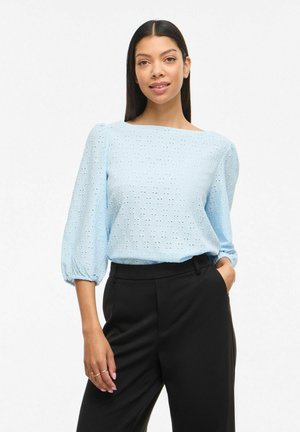 Kvinne med rett mørkt hår iført en lyseblå bluse med hullbroderi og puffede ermer, og svarte bukser med høyt liv, med en hånd i lommen.