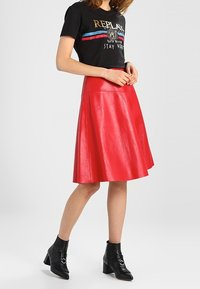 Woman wearing a black graphic t-shirt, red A-line leather skirt, and black ankle boots with buckles, standing against a plain background.