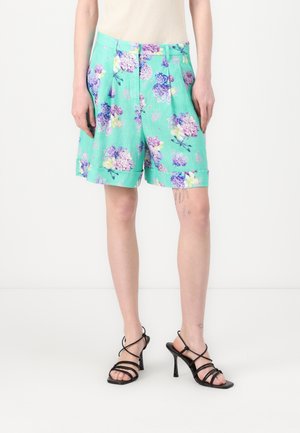 Grønne blomstrede shorts med lilla og gule blomster, plisseret design, sidelommer og optrullede manchetter. Båret med sorte stroppede højhælede sko.