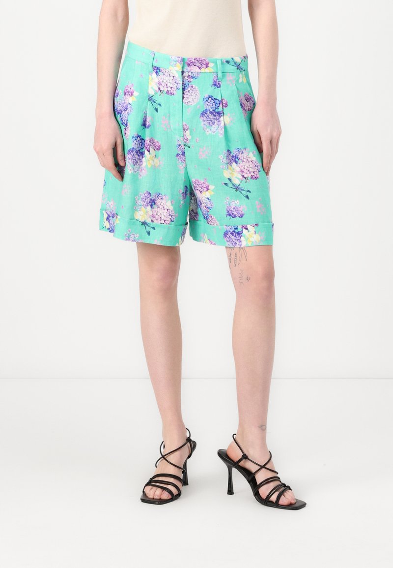 Grønne blomstrede shorts med lilla og gule blomster, plisseret design, sidelommer og optrullede manchetter. Båret med sorte stroppede højhælede sko.