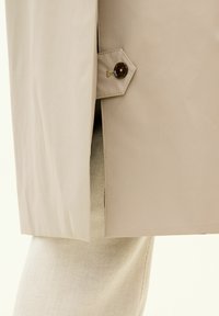 Dettaglio di un impermeabile beige che mostra una chiusura laterale con bottone lucido di colore marrone e una fodera texturizzata di pantaloni in corduroy chiaro.