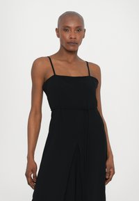 Black sleeveless dress with a square neckline, thin straps, and a tied waist. Features a front slit design for added movement.