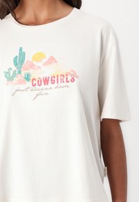 Cream-colored cotton t-shirt with a crew neck, featuring a graphic design of cacti, mountains, and the text "COWGIRLS just wanna have fun."