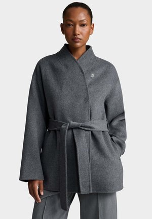 Donna che indossa un cappotto di lana grigio con cintura, maniche larghe e collo alto, in piedi con una mano in tasca su sfondo semplice.