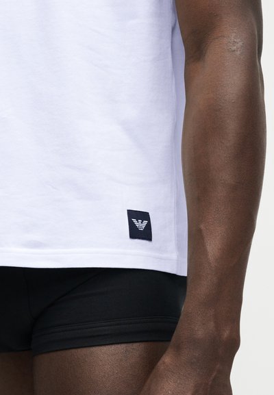 Primo piano di una persona che indossa una maglietta bianca con il logo Emporio Armani vicino all'orlo e pantaloncini neri.