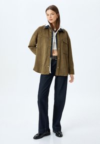 Giacca in suede verde oliva con collo e tasca frontale, abbinata a pantaloni in denim scuri a vita alta e scarpe nere.