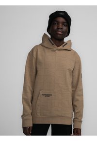 Sudadera beige con un patrón texturado de letras, bolsillo frontal y acentos negros en la capucha. Combinada con un gorro negro.