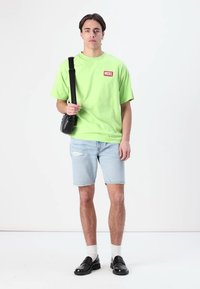 Neonově zelené oversized tričko s červeným logem, světle modré džíny šortky, bílé ponožky a černé polobotky. Model stojí na bílém pozadí.