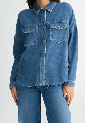 Femme portant une veste en jean bleue avec des fermetures à boutons et des poches à rabat sur le devant, assortie à un jean bleu.