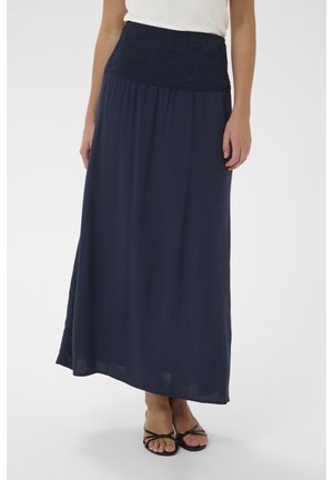 Kvinde iført en navyblå maxinederdel med smocket talje og sorte sandaler med stropper, stående mod en hvid baggrund.