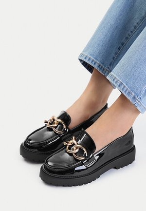 Mocasines negros brillantes con suelas gruesas y gran cadena de oro decorativa, usados con vaqueros azul claro recortados y pies descalzos.