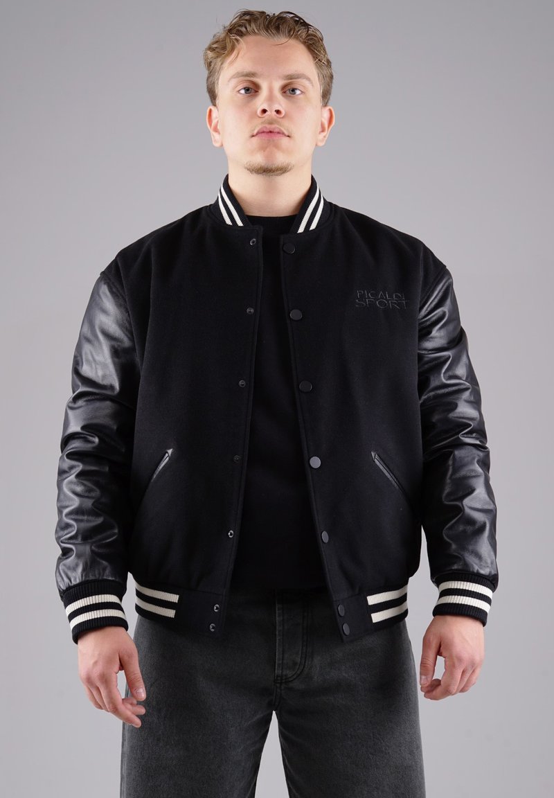 Schwarze Collegejacke im Varsity-Stil mit Wollkörper, Lederärmeln, gestreiften Bündchen und einer Vorderinnentasche. Enthält ein dezentes Logo auf der Brust.
