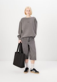 Felpa grigia oversize con logo stampato, pantaloni shorts in denim grigi, borsa a tracolla nera, scarpe da ginnastica nere con strisce bianche e calzini bianchi a caviglia.