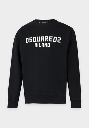 Jeune homme portant un sweat-shirt noir Dsquared2 Milano, un short noir, des chaussettes blanches et des baskets noires, debout devant un fond uni.