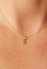 Collier en chaîne dorée avec pendentif en forme de "r" minuscule, texture lisse avec finition brillante, reposant sur une peau nue.