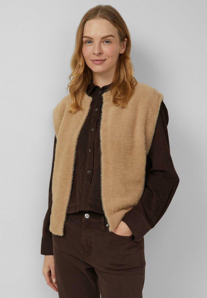 Beige mouwloos vest van zacht pluche stof, met een zachte textuur, open voorkant en een contrasterend donker kledingstuk eronder.