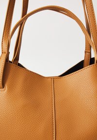 Torebka tote w kolorze brązowym z teksturowanym wykończeniem, wyposażona w dwie mocne rączki i strukturalny design. Otwarta górna część z czarnym wnętrzem.
