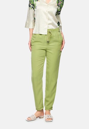 Woman wearing light green drawstring pants, white patterned button-up shirt, and white woven slide sandals on a white background.