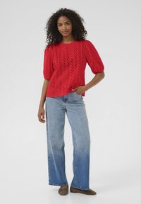 Rode, kort-één mouw gebreide top met een gestructureerd chevronpatroon, gecombineerd met lichtblauwe wijde jeans en bruine ballerina's. Model staat rechtop, met het gezicht naar voren.