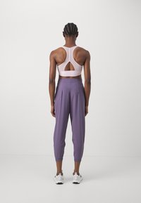 Jasnoróżowy stanik sportowy z wycięciem, zestawiony z luźnymi, zwężającymi się lawendowymi spodniami. Widoczne białe buty sportowe. Minimalistyczny design.