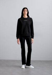 Μαύρο φούτερ με στρογγυλή λαιμόκοψη και κεντημένο κείμενο, σε συνδυασμό με μαύρα εφαρμοστά παντελόνια και λευκά αθλητικά παπούτσια, σε ουδέτερο φόντο.