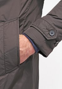 Tmavě šedý prošívaný rukáv bundy, manžeta se zapínáním na dva knoflíky. Kapsa na boku s viditelným prošitím. Vrstvené s tmavě modrou košilí pod ním.