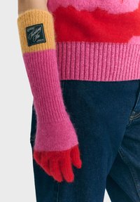 Hånd iført en lang strikket handske med gule, pink og røde farveblokke og et sort mærke, parret med en pink og rød sweater samt mørke jeans.