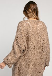 Wełniany kardigan w jasnobeżowym kolorze o luźnym fasonie, prezentujący skomplikowany wzór koronkowy oraz szerokie rękawy. Widoczna wyraźna faktura dzianiny.