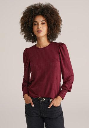 Haut long à manches maroon en tissu texturé, col rond et manches bouffantes. Associé à un jean noir taille haute et une ceinture dorée.