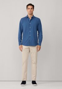 Camicia blu a maniche lunghe con bottoni, tessuto morbido, vestibilità regolare, abbinata a pantaloni chino beige e sneakers nere, in piedi davanti a uno sfondo grigio chiaro.