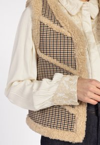Taan- en zwart geruit vest met een fluffy beige rand, over een witte blouse met bloemendecoraties op de mouwen.