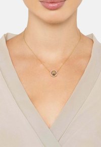 Collar de oro con un colgante circular que presenta un diseño de abeja, rodeado de pequeñas piedras transparentes, llevado sobre una blusa de color claro.