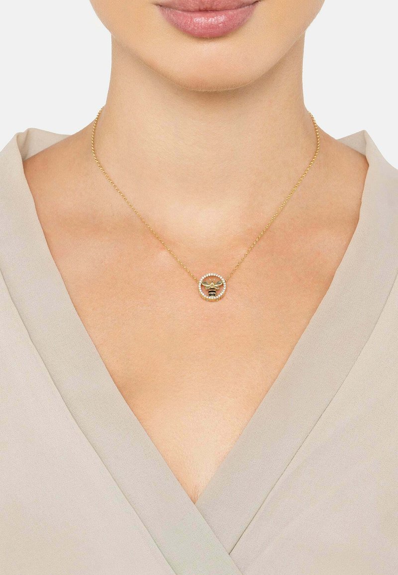 Collar de oro con un colgante circular que presenta un diseño de abeja, rodeado de pequeñas piedras transparentes, llevado sobre una blusa de color claro.