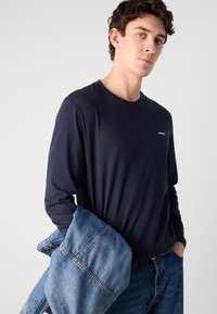 Navy long-sleeve t-shirt made of soft fabric, featuring a round neck and a subtle logo. Model holds a light blue denim jacket.