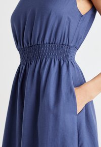 Navy blue sleeveless dress with a smocked waist, soft texture, and side pockets; flowing A-line shape with gathered fabric.