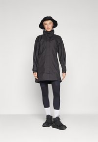 Black waterproof jacket with a high collar, long sleeves, and zip closure, paired with black leggings, white socks, and a black bucket hat.