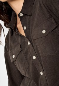 Camisa de pana marrón con cuello puntiagudo, que cuenta con dos bolsillos en el pecho y botones de color claro, destacando un diseño texturizado.