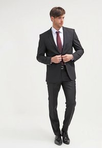 Zwart op maat gemaakt pak met een insnijding revers, wit overhemd en een bordeauxrode stropdas met polkadots. Soepele textuur, nauwsluitende pasvorm, gecombineerd met glanzende zwarte schoenen.