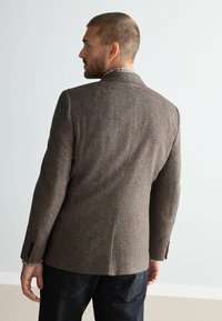 Brown herringbone jacket with notched lapels, two back vents, and buttoned sleeves; worn over dark denim pants, showcasing a tailored fit.