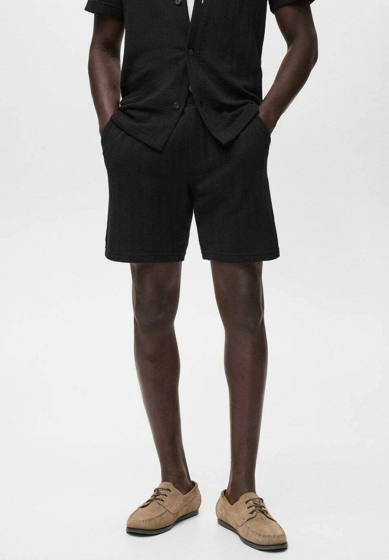 Homme portant un short texturé noir et une chemise à manches courtes assortie, avec des chaussures à lacets en suède beige, les mains dans les poches, se tenant sur un fond blanc.