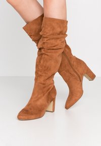 Botas de ante marrón de media caña con un diseño holgado, tacones de bloque y cremalleras laterales, llevadas con las piernas cruzadas sobre un fondo blanco liso.