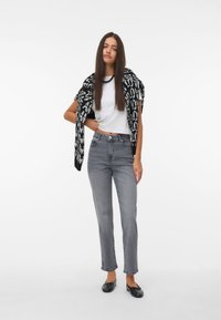 Bela majica, šal s črno-belim vzorcem, sive hlače ravnega kroja in črne ravne čevlje. Manekenka stoji z eno roko na boku.