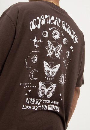 Brown t-shirt with white mystical designs including butterflies, moon phases, eyes, sun, stars, and text "Mystical Rising" and "Live by the Sun, Live by the Moon."